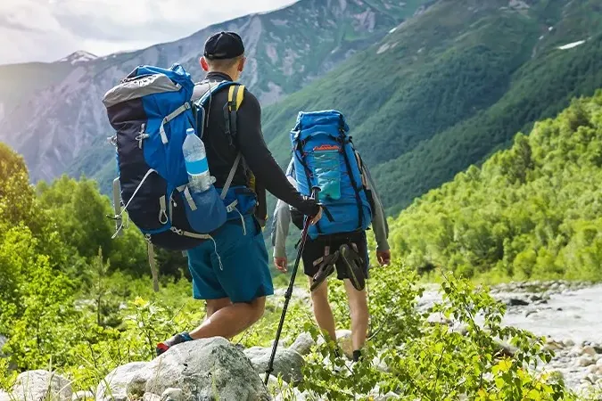 Doi drumeți cu rucsacuri mari și bețe de trekking pe o potecă de munte, lângă un pârâu
